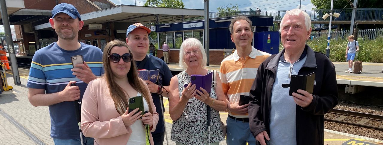 Members of Bedfordshire SLC are stood with Samantha Leftwich, Engagement Manager for East England. They are stood on a platform at Stevenage train station as part of the Aira trial. They are all holding their smartphones in one hand, their long cane in the other.
