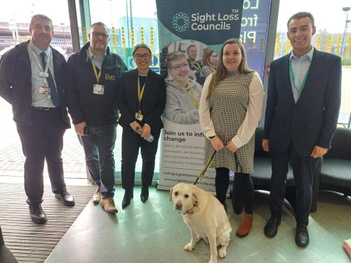 Five people in a group photo, including Engagement Manager Sam and GTR staff members, at the Bedfordshire 'Coffee and Chat' event at Luton Airport Parkway with the SLC banner visible behind them.