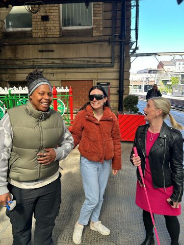 Three women at a train station. From left to right: a woman wearing a green gilet is guiding the woman standing beside her, who is wearing sim specs. Sight Loss Council Engagement Manager Kelly is standing on the right talking to them.