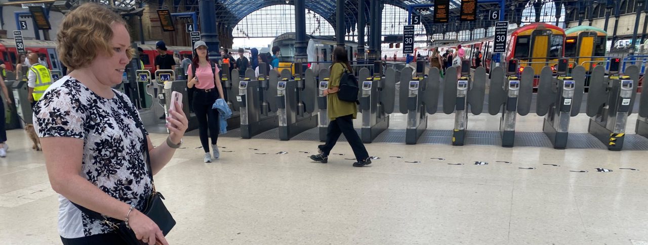 SLC volunteer Linn walking through a train station using the Aira app. She has her smartphone in one hand and her white navigation cane in the other.