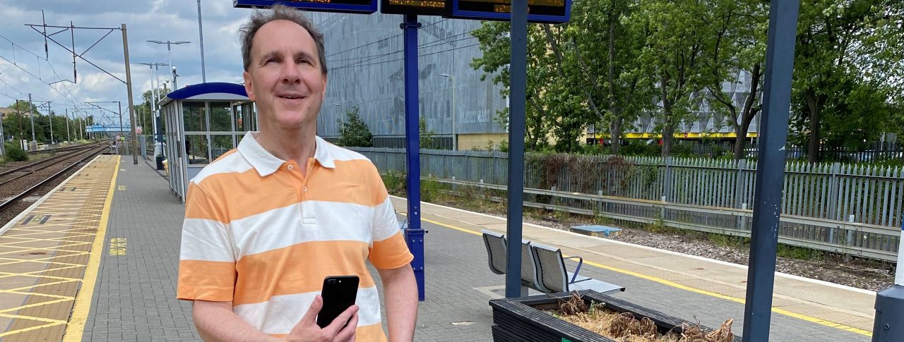 Paul Day, Bedfordshire SLC member, is standing on a platform at Stevenage train station. he is holding his long cane in one hand, and his phone up in the other.