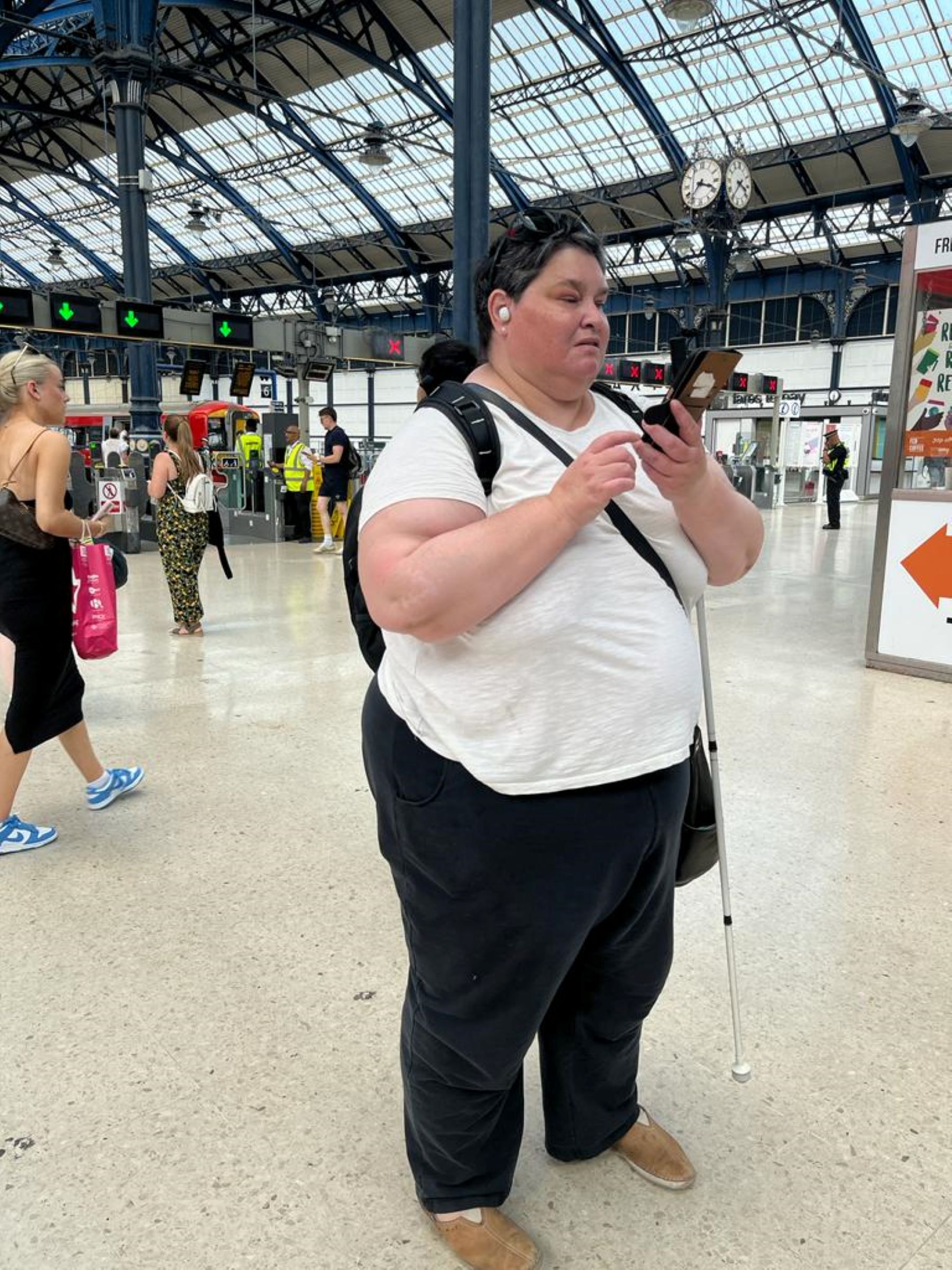 Sussex SLC member Iris Keppler using the AIRA app to navigate around Brighton train station. Iris is pictured standing in the middle of a busy station, with the platforms to her left and lots of people walking behind her.