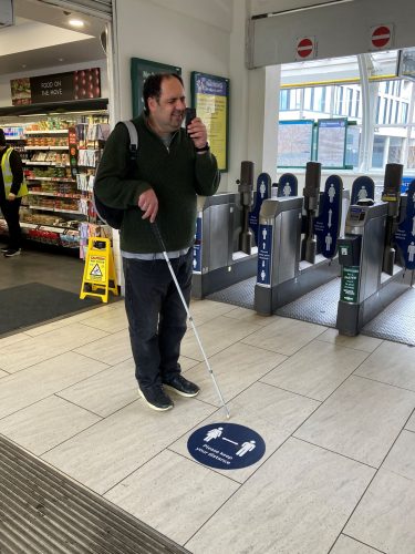Perrie, Sutton Vision Rep navigating a train station using his white cane while testing the AIRA app. Perrie is pictured with two train station barriers to his right and a supermarket behind him.