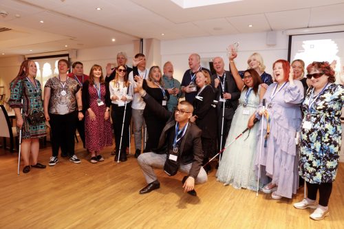 A group photo of eighteen people celebrating. They are smiling and cheering and many are wearing medals, with a few people also holding their long canes. They are all smartly dressed and look happy. One man is crouched at the front with his arm in the air, celebrating.