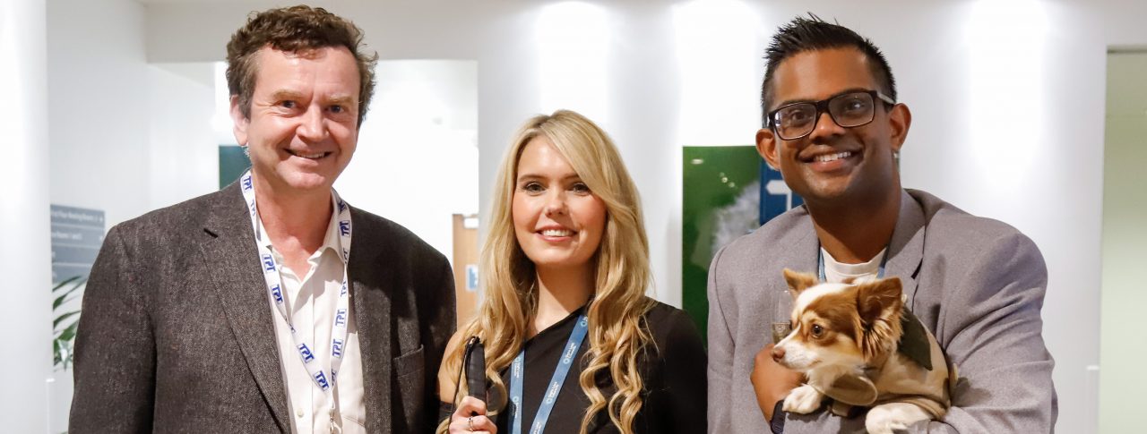 This photo is of CEO Charles Colquhoun, Claire Sisk, and her male colleague. They are all dressed smartly and smiling at the camera. Charles is on the left, and is wearing a dark grey suit jacket, a white shirt, brown trousers, and has brown hair. Claire is in the middle, wearing a black dress and holding her long cane in her right hand. She has a small pink handbag over her left wrist, and is holding hands with her colleague, who is on the right of the photo. He is wearing a light grey suit jacket, a white top, black trousers, and has dark brown hair and glasses. He is also carrying a small brown-and-white chihuahua.