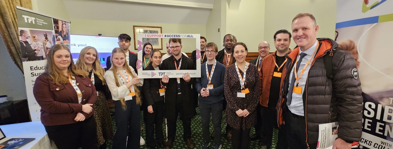 A group of around 15 people stand closely together indoors, smiling at the camera. In the centre, two people are holding a large cardboard photo frame that reads 'I support #ACCESSFORALL'. The Thomas Pocklington Trust logo and website are clearly visible at the bottom of the frame.