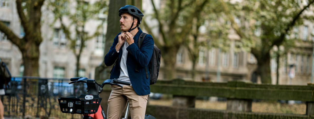 White man fastens a black helmet while sitting on an orange Voi e-bike, preparing to cycle off. He is smartly dressed in beige chinos and a navy jacket.