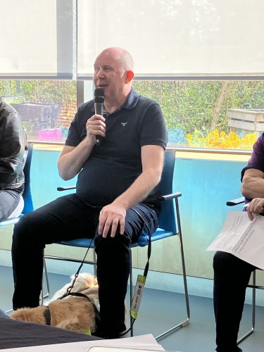 BaBC SLC member Paul Hopkins sitting on a chair and talking into a microphone. Paul is wearing a navy coloured polo shirt and dark trousers.