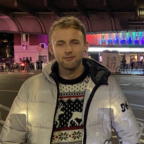 Student Voices volunteer, Aleks, is smiling at the camera. He is wearing a Christmas jumper and a white coat.