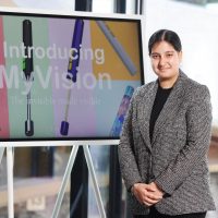 Student Voices volunteer, Ramneek, is looking towards the camera and smiling. She is standing in front of a large presentation screen that reads 'Introducing MrVision'. Ramneek is wearing a smart grey blazer and has her hands clasped together in front of her.