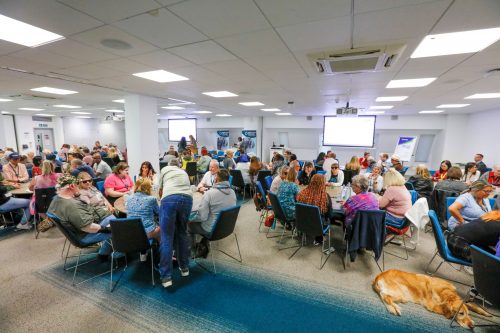 Sight Loss Council conference delegates (SLC volunteers) and sighted guides sat around round tables int he main conference hall. SLC banners are in the background, flip chart two larges screens to show the presentations. A golden coloured guide dog is laid on the floor.