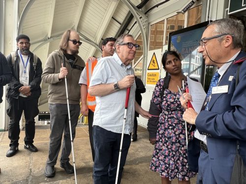 Antony Merlyn, Accessibility Engagement Manager at GTR, talking to attendees at Sutton. There are six people, three of whom are holding long canes. Antony is wearing a smart navy suit, with a striped, white shirt, and blue tie. He is a middle-aged man and is wearing glasses, and is his hands he is holding a pen and red notebook. He is speaking to four men and one woman.