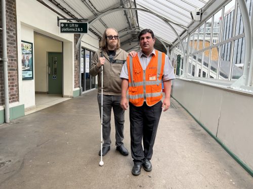 Attendee Gus is being guided by Noor at Sutton railway station. Gus is on the left, wearing black glasses and holding a long cane. He is wearing black shoes, black, denim jeans, and a brown zip-up hoodie. He has shoulder-length brown hair and a beard. Noor is on the right and is guiding Gus through the station. Noor is wearing his uniform, which is black shoes, smart black trousers, a light-coloured smart shirt, and an orange high-visibility jacket with his name badge on it. He has short, dark hair. They are both facing the camera.