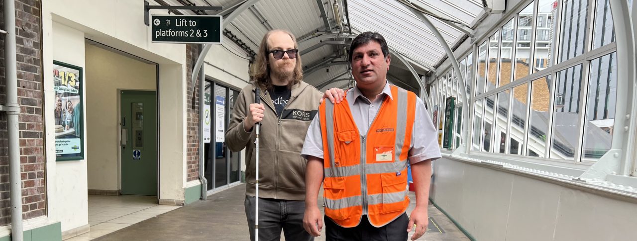 Attendee Gus is being guided by Noor at Sutton railway station. Gus is on the left, wearing black glasses and holding a long cane. He is wearing black shoes, black, denim jeans, and a brown zip-up hoodie. He has shoulder-length brown hair and a beard. Noor is on the right and is guiding Gus through the station. Noor is wearing his uniform, which is black shoes, smart black trousers, a light-coloured smart shirt, and an orange high-visibility jacket with his name badge on it. He has short, dark hair. They are both facing the camera.