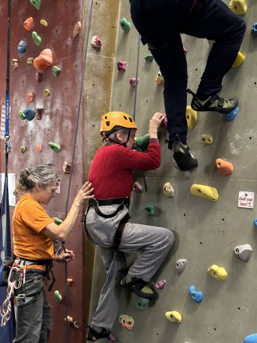 Two people wearing colourful helmets climbing the climbing wall at a previous multi-sport taster event. A third lady, wearing a yellow sweat-shirt, stands at the bottom to support the climbers.