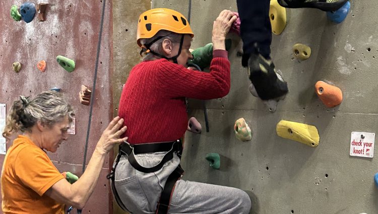 Two people wearing colourful helmets climbing the climbing wall at a previous 'Let's Get Active' event. A third lady, wearing a yellow sweat-shirt, stands at the bottom to support the climbers.