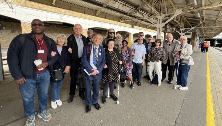 A group shot of 17 people of varying ages, genders, and ethnicities standing together at Wimbledon station. Five people are holding their long canes. Some people are carrying rucksacks, handbags, and coffee cups. Antony Merlyn stands at the front of the group in the middle, smiling at the camera. He is smartly dressed in a navy suit and tie, is holding a notebook, and carrying a rucksack.