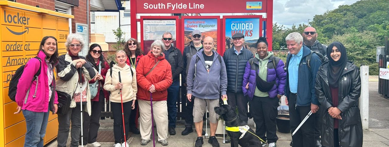 A group of seven people of mixed ages, genders and ethnicities. Some are holding white canes and there is one person with a guide dog. The group is standing in front of a red sign that reads, 'South Fylde Line' at a train station. There is an Amazon locker to the left and trees in the background.