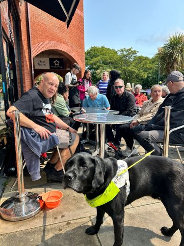 A group of five people are seated around a metal table outside a brick building with a sign that reads 'Hey Treat.' The people are engaged in conversation and appear to be enjoying their time together. In the foreground, there is a black guide dog wearing a yellow harness standing on the pavement next to an orange water bowl.
