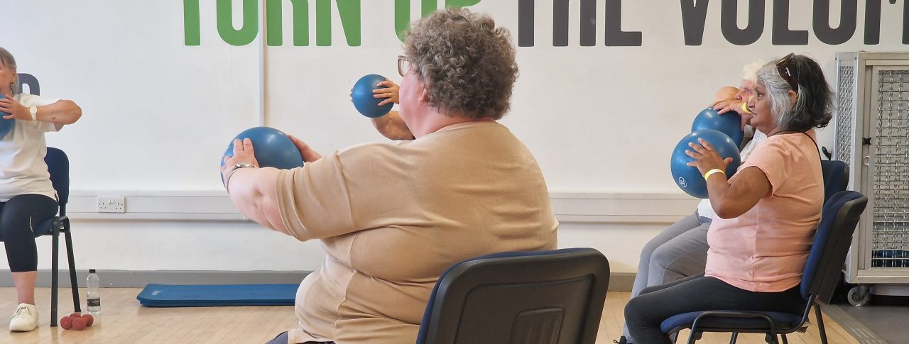 Five delegates sitting on chairs inside a sports hall, with the three at the front most visible. They are holding blue balls, which they are using to exercise. They are dressed in comfortable clothing, and there are water bottles, and dumbbells on the floor by their chairs. It says 'Turn up the volume' in big green and black capital letters on the back wall.