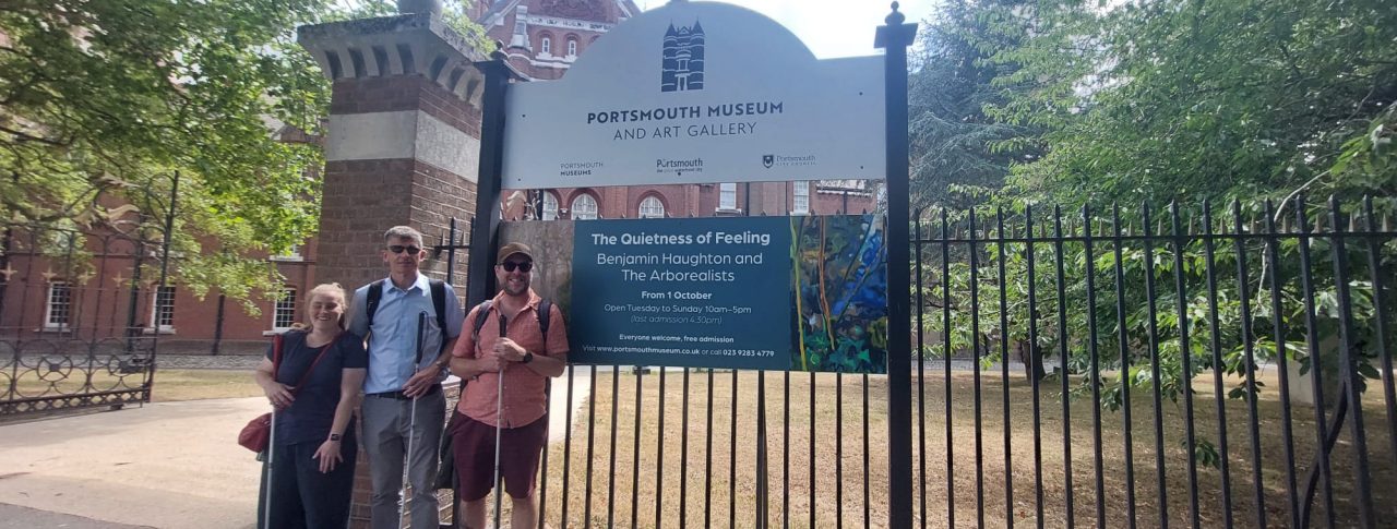 Three people, a woman and two men, are standing slightly to the left of a sign in front of a large building. The people are all holding white walking sticks, and the two men in the centre and to the right are wearing visors. The sign the people are standing next to says ‘Portsmouth Museum and Art Gallery” while an additional sign below is promoting an event for, ‘The Quietness of Feeling’ on the 1st of October.