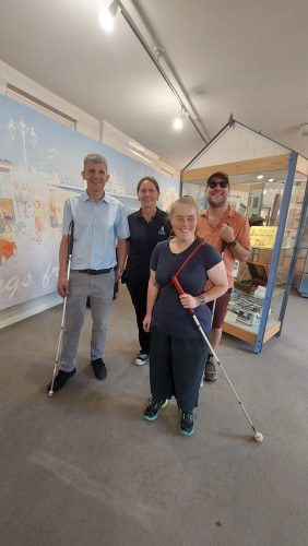 A group of four people stood inside Portsmouth Museum and Art Gallery. Three of the people are standing next to a staff member of the museum alongside a mural in the background. A cabinet displaying historical items is behind the group.