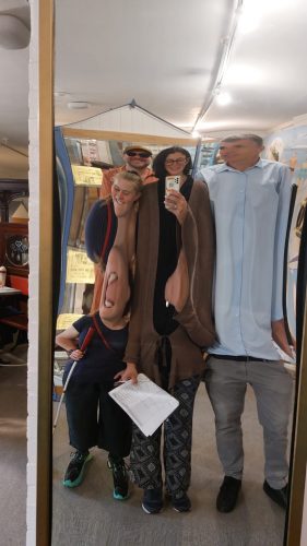 A group of people are taking a photo in front of a distorted mirror in at Portsmouth Museum. The group are all smiling, as their bodies are under the illusion of being stretched from their neck to their torsos.