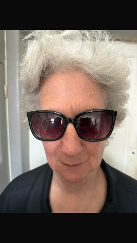 Steph Syson headshot. Steph has shot curly grey hair and is wearing sunglasses and a black t-shirt. She is smiling at the camera. 