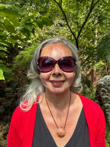 Patricia Clements headshot. Patricia has long light hair and is wearing a red cardigan and black top with a statement necklace and sunglasses. She is smiling at the camera.