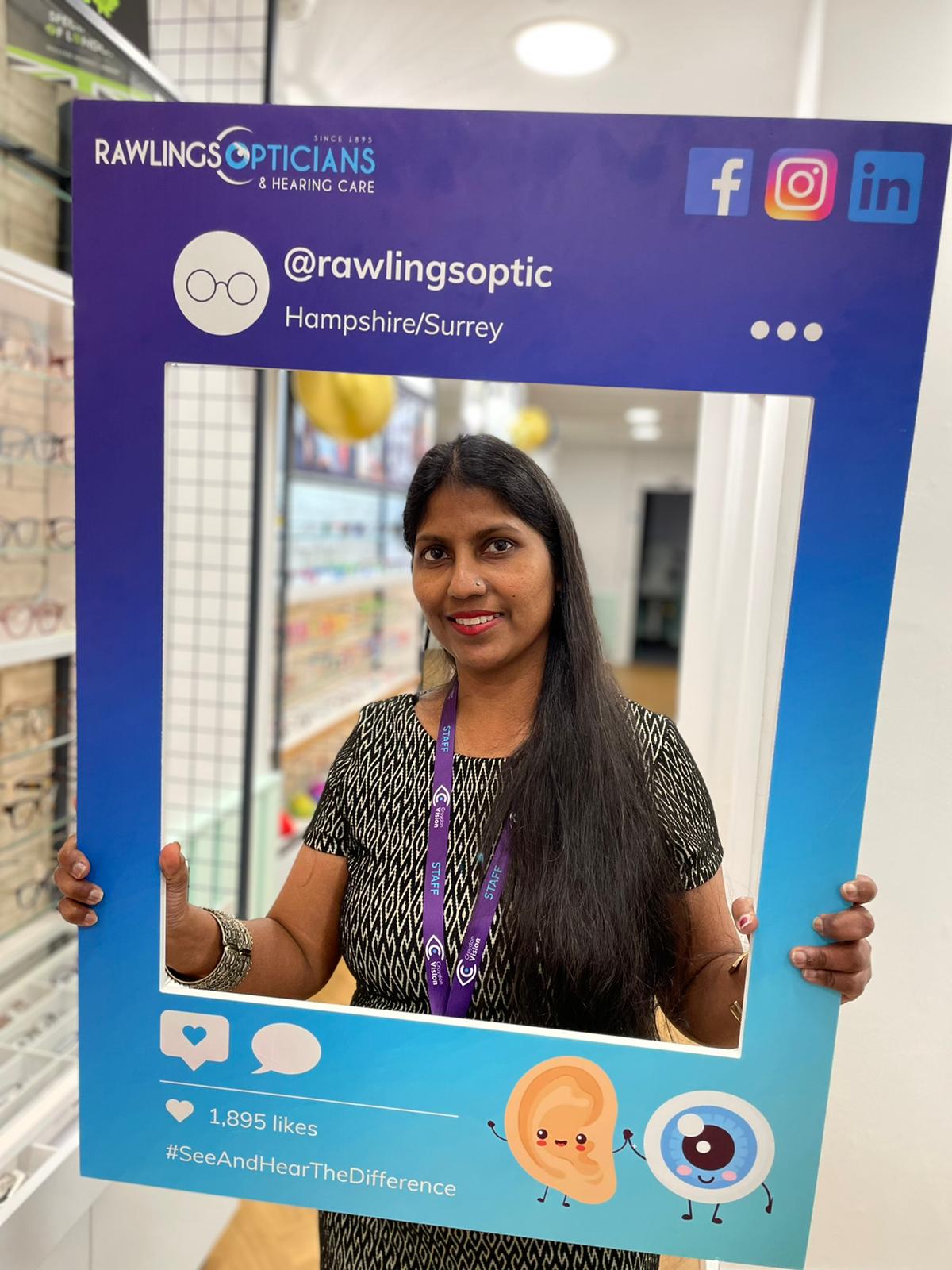 Shalini stands inside an optician's store, holding a large promotional frame in blue and purple colours. The frame features branding for 'Rawlings Opticians & Hearing Care' and includes social media handles, hashtags, and cartoon icons of an ear and an eye.