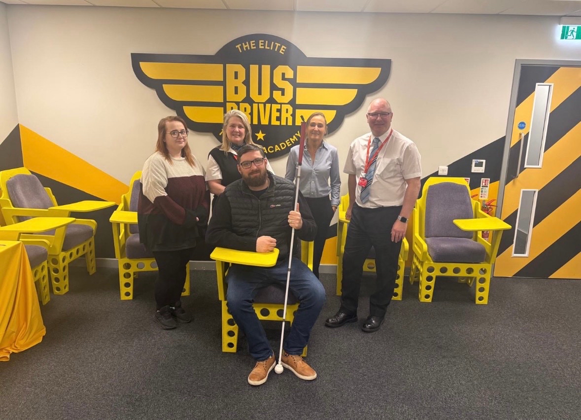 A group of five people are standing and smiling at the camera in a bright, modern training room. The setting is the Go North East “Bus Driver Academy”, as indicated by the bold yellow and black signage on the wall behind them. Jack Moffat (north engagement manager for TPT)in the center is seated and holding a white cane, highlighting their lived experience of sight loss. They are surrounded by four colleagues standing on either side. The chairs in the room are painted bright yellow with integrated writing desks, giving the space a training or workshop feel. The atmosphere is professional and positive.