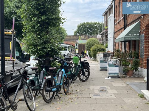 A pavement is cluttered up with multiple bikes that have not been parked to the edge to allow pedestrians to walk freely. A narrow gap is present making navigating a struggle for BPS people.