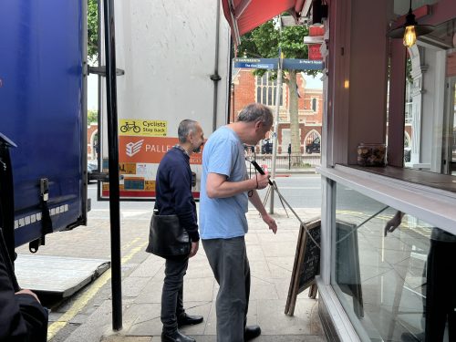 Two men, including SLC co-ordinator Liam, are trying to navigate their way on some pavement as a lorry's backdoors are open and cover a large portion of the pavement, making their ability to walk along it difficult. Liam is on the right holding a white-cane while trying to get through the narrow gap.