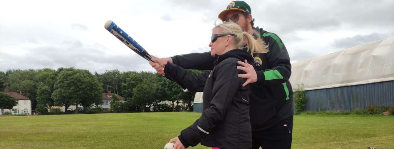 Kelly Barton, Engagement Manager for the North West during a VI Baseball session. She is standing on the field at the home base. She is with an instructor who is guiding her where to position the bat. Kelly is holding the baseball in her other hand.