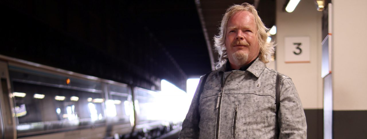 Portrait image of Steve Keith, Birmingham and Black Country SLC member, standing on a train platform with his guide dog. A train is on the opposite side of the tracks.