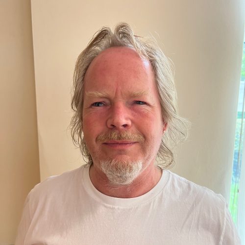 Headshot of Sight Loss Council volunteer Steve. He has antique blonde hair, a moustache, and a short beard on his chin. He is wearing a white t-shirt and smiling at the camera.