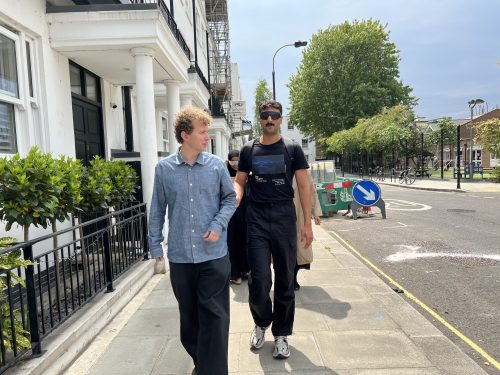 Two men are walking along a pavement. Theo on the left and is supporting Amric who is wearing Sim-Specs.
