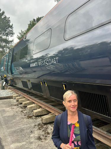 SLC North West Engagement Manager, Kelly stands beside a disused Avanti West Coast train, whilst holding a pink-cane.