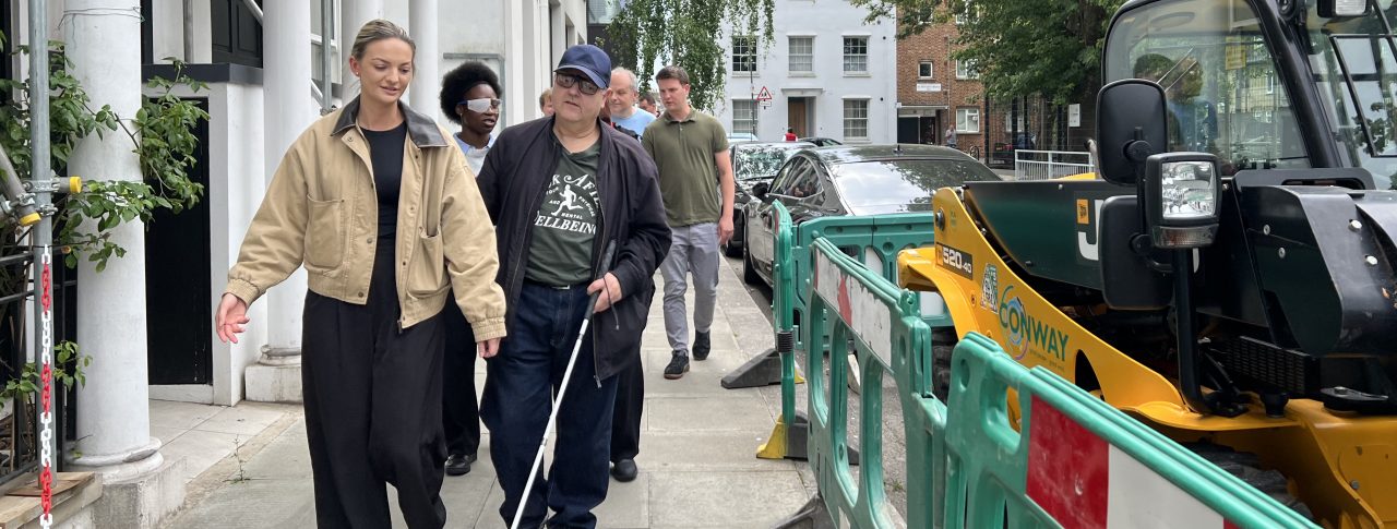 People are walking along a path with a woman in a beige jacket and a man using a white-cane to help navigate his way. The path is narrow due to some machinery and fencing to the right of the image.