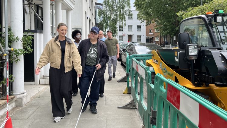 People are walking along a path with a woman in a beige jacket and a man using a white-cane to help navigate his way. The path is narrow due to some machinery and fencing to the right of the image.