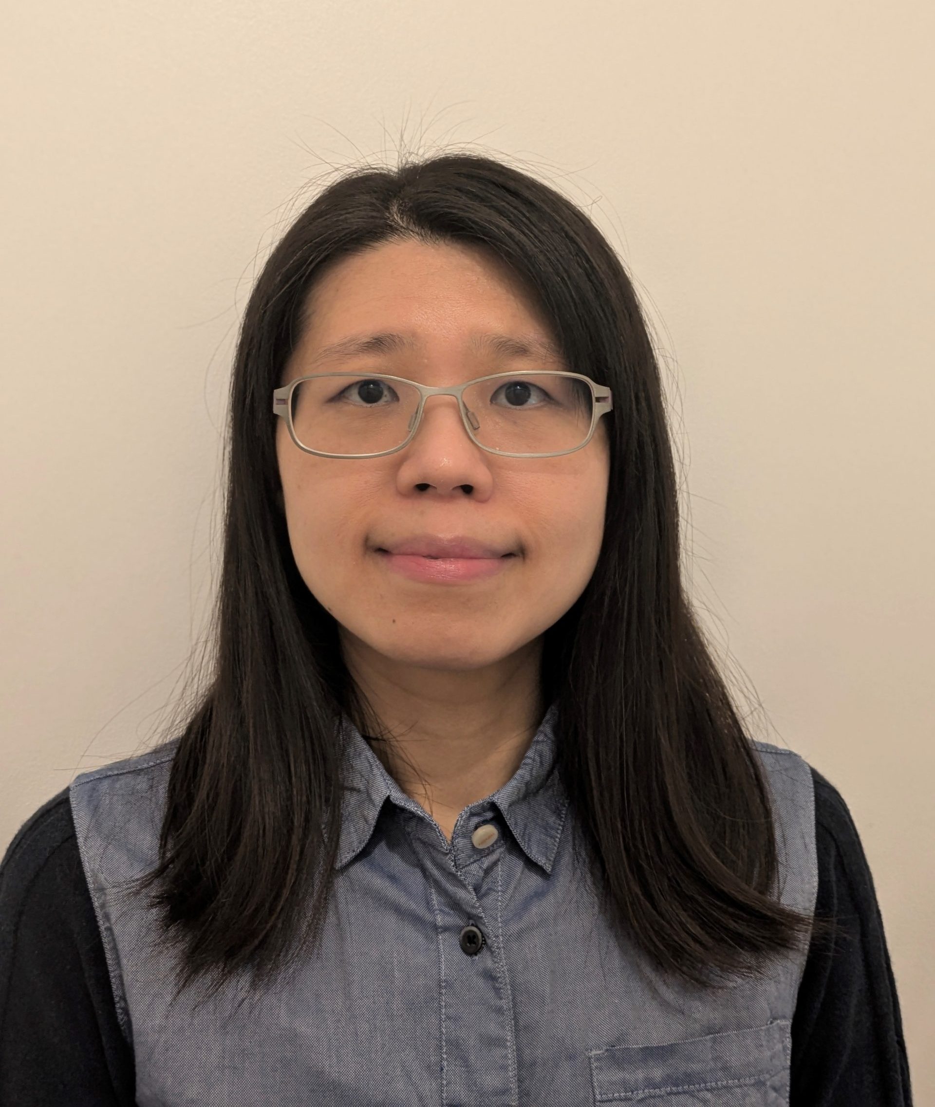 Head shot of Ching, SE London SLC volunteer. Cheer is standing against a beige wall, wearing clear framed glasses, smiling at the camera.