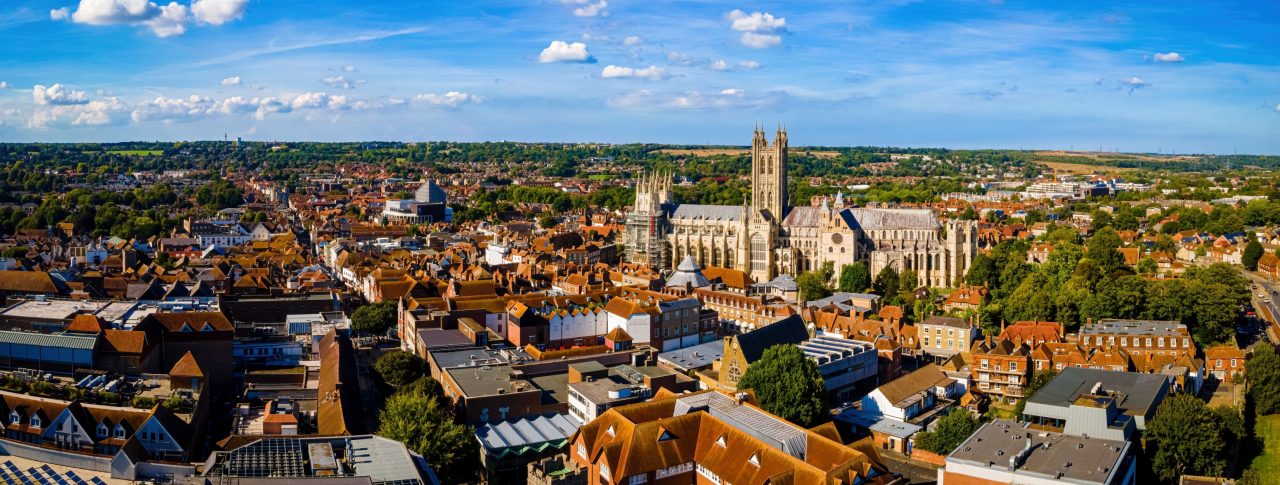 Aerial view of Canterbury, cathedral city in southeast England. The cathedral can be seen in the centre of the shot to the right, surrounded by old buildings and greenery. A blue sky with scattered clouds.