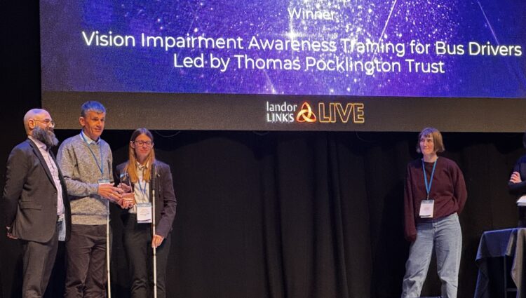 On stage at the award ceremony, a rather snazzy looking Dave and Lauren stand, holding their long canes next to a gentleman in a dark grey formal suit sporting a wonderfully bushy beard. A large screen behind them informs us of the celebrated winners with text that reads: Bus Centre of Excellence, Awards 2025, Collaboration Award, Winner, Vision Impairment Awareness Training for Bus Drivers Led by Thomas Pocklington Trust. The text is white on a sparkly blue background. The Screen behind is at least two Daves tall, and the stage must be at least four Daves wide, and the trio are standing to the left of the stage. To the right are two people who I am assuming are facilitators, one of whom is standing on a table draped in black cloth and the other standing looking towards the trio in awe. The lighting is an ambient blue with a soft spotlight on the winners.