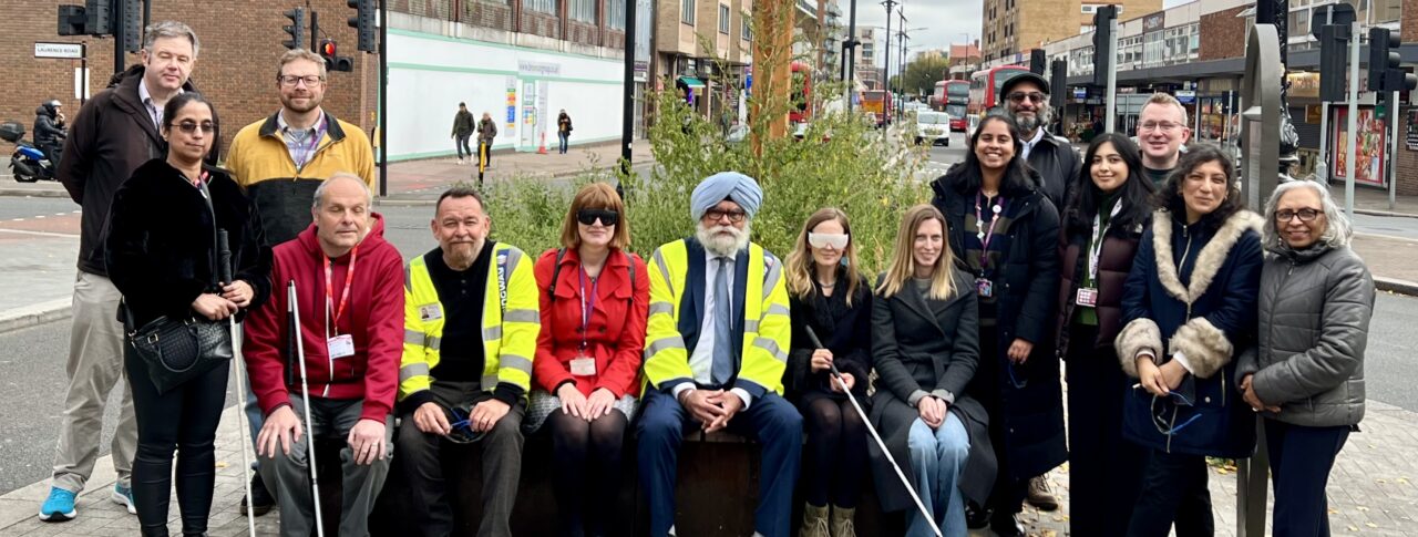 A group of 15 people including representatives from Hounslow Council and Sight Loss Council volunteer members gathered on a pavement in Hounslow Town Centre near a planter, smiling at the camera. Some are seated on a bench, others standing, and several hold white canes. Two individuals from Hounslow Council wear high-visibility jackets. Urban buildings, traffic lights, and a cloudy sky form the backdrop.