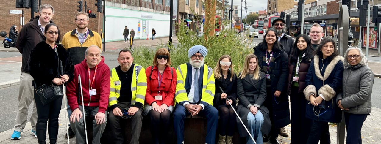 A group of 15 people including representatives from Hounslow Council and Sight Loss Council volunteer members gathered on a pavement in Hounslow Town Centre near a planter, smiling at the camera. Some are seated on a bench, others standing, and several hold white canes. Two individuals from Hounslow Council wear high-visibility jackets. Urban buildings, traffic lights, and a cloudy sky form the backdrop.