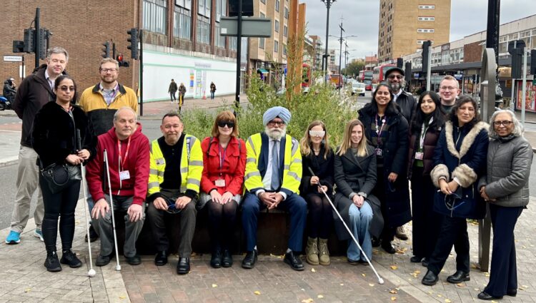 A group of 15 people including representatives from Hounslow Council and Sight Loss Council volunteer members gathered on a pavement in Hounslow Town Centre near a planter, smiling at the camera. Some are seated on a bench, others standing, and several hold white canes. Two individuals from Hounslow Council wear high-visibility jackets. Urban buildings, traffic lights, and a cloudy sky form the backdrop.