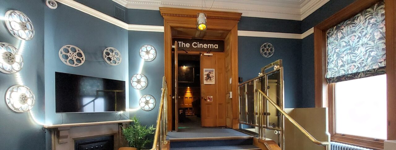 Entrance area to Guildhall Cinema with blue walls decorated with film reels, a flat-screen TV, and a small seating area. A staircase with brass handrails leads up to a wooden doorway labelled “The Cinema.” A patterned blind covers a large window on the right.