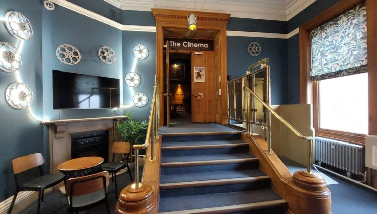 Entrance area to Guildhall Cinema with blue walls decorated with film reels, a flat-screen TV, and a small seating area. A staircase with brass handrails leads up to a wooden doorway labelled “The Cinema.” A patterned blind covers a large window on the right.