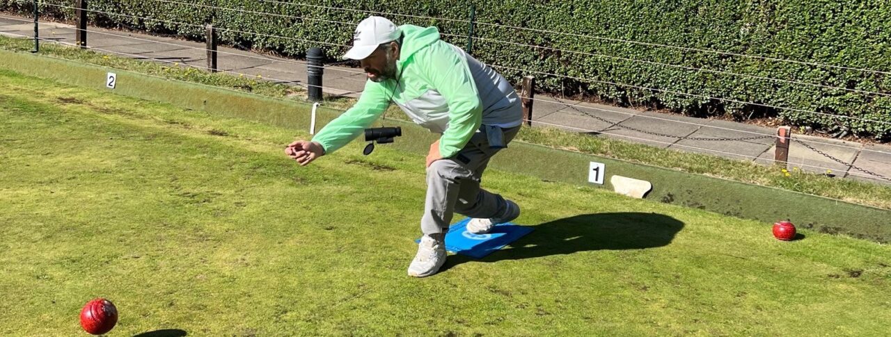 Portrait image of SE London volunteer, Steve, playing bowls on a bowling lawn. He is crouching down, having bowled his ball.
