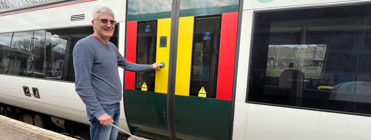 A male wearing dark glasses is standing on a train station platform, holding a white cane and pressing the door button on a train before he boards. The train door is marked with bright yellow and red panels for visibility, and symbols above the windows indicate spaces for wheelchair users and bicycles. Trees and a blue sky are visible in the background.